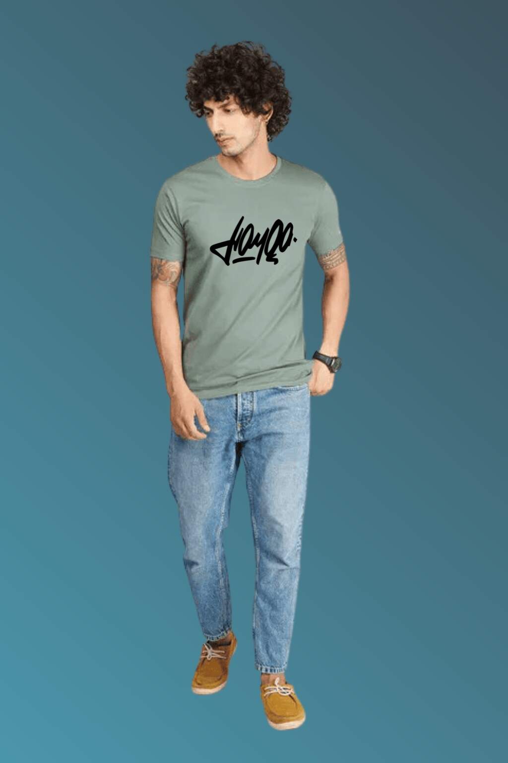 t-shirt featuring bold black stylized typography, worn with blue denim jeans and brown casual shoes. Set against a smooth blue gradient backdrop, the photo highlights modern streetwear style in a minimal, fashion-forward composition."